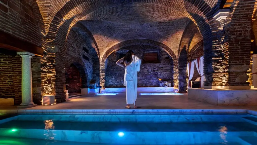 An elegant woman in a luxurious dressing gown stands in front of the main pool, in a sophisticated setting that emphasises the architecture of In Acqua Veritas